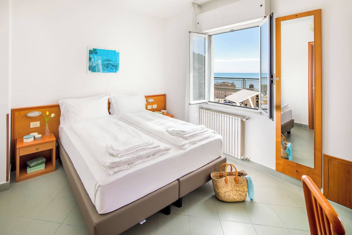 A hotel room with a bed, a window with a view, a bedside table, a nightstand, a mirror, and a wall-mounted picture.