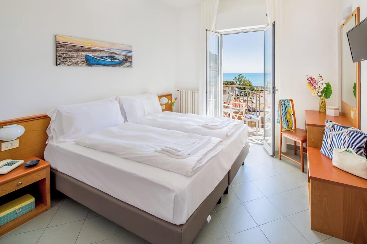 A hotel room with a bed, a desk, a chair, a TV, and a balcony with a view of the sea.