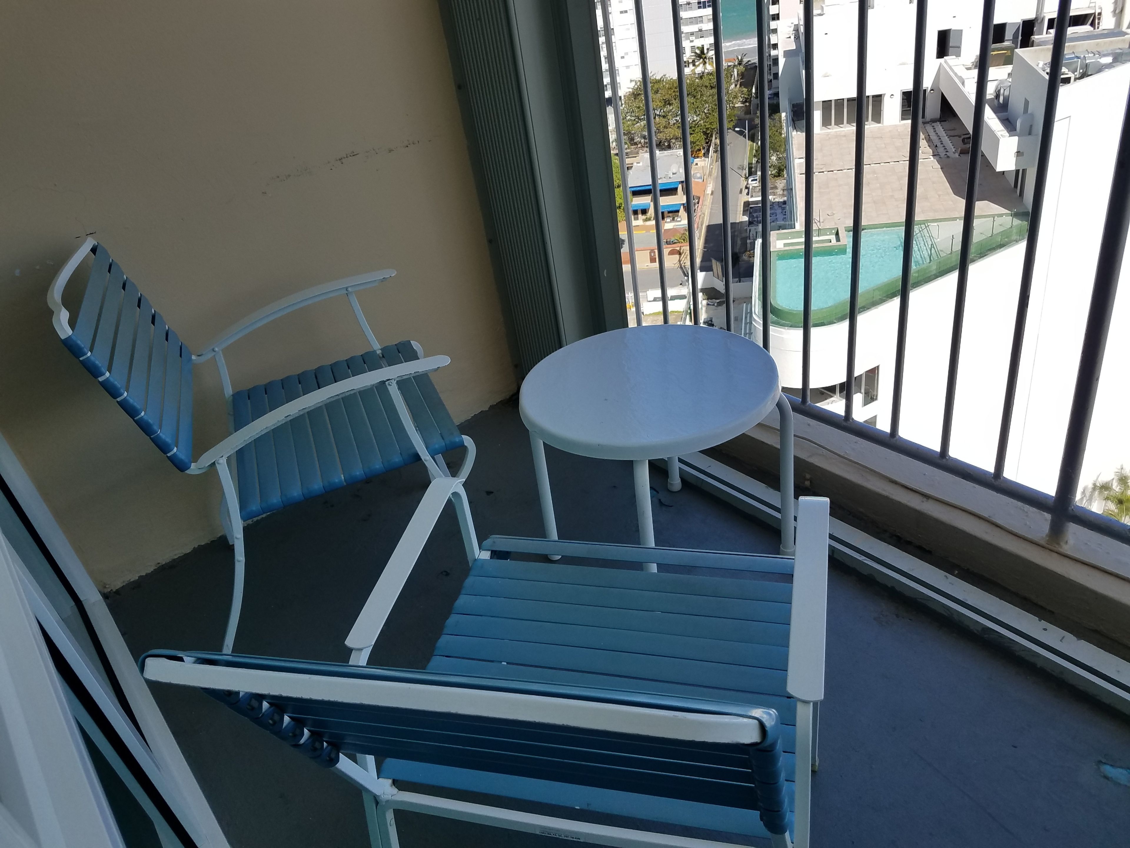 Un balcón con una silla azul, una mesita redonda y vistas a una piscina y edificios.