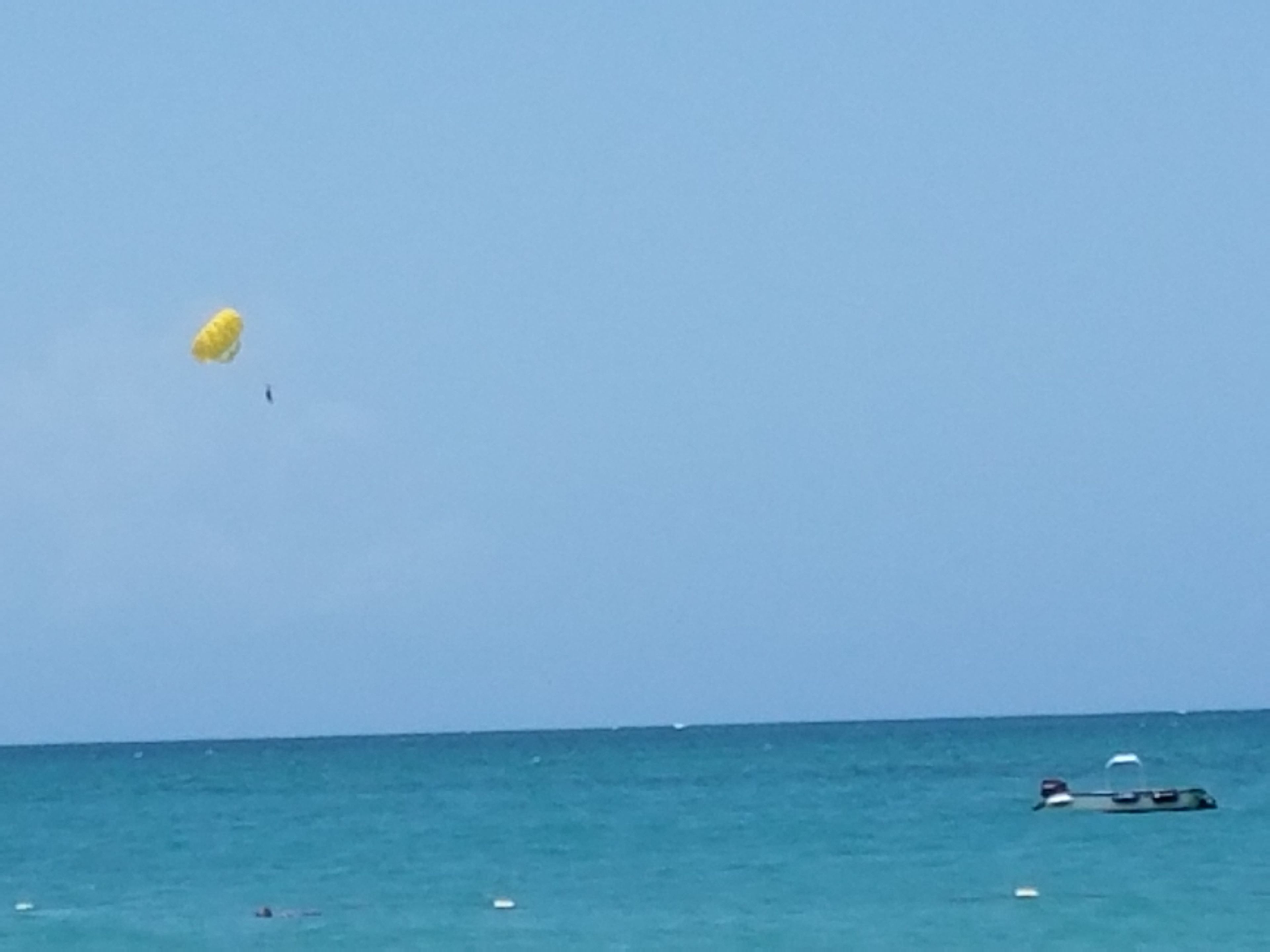 Un ciel bleu et dégagé surplombe une mer calme, avec un parachute ascensionnel jaune et un petit bateau à l'horizon.