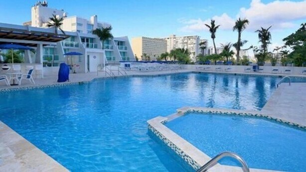 A swimming pool with lounge chairs and a covered seating area, surrounded by palm trees and modern buildings. - Mare 1660 (Carolina)