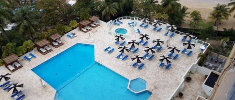 A rooftop pool with blue water, surrounded by lounge chairs and umbrellas, overlooking a beach with palm trees and the ocean.