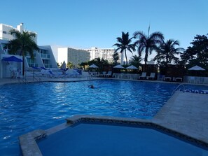 A swimming pool with lounge chairs and umbrellas, surrounded by palm trees and modern buildings. - Mare 1660 (Carolina)