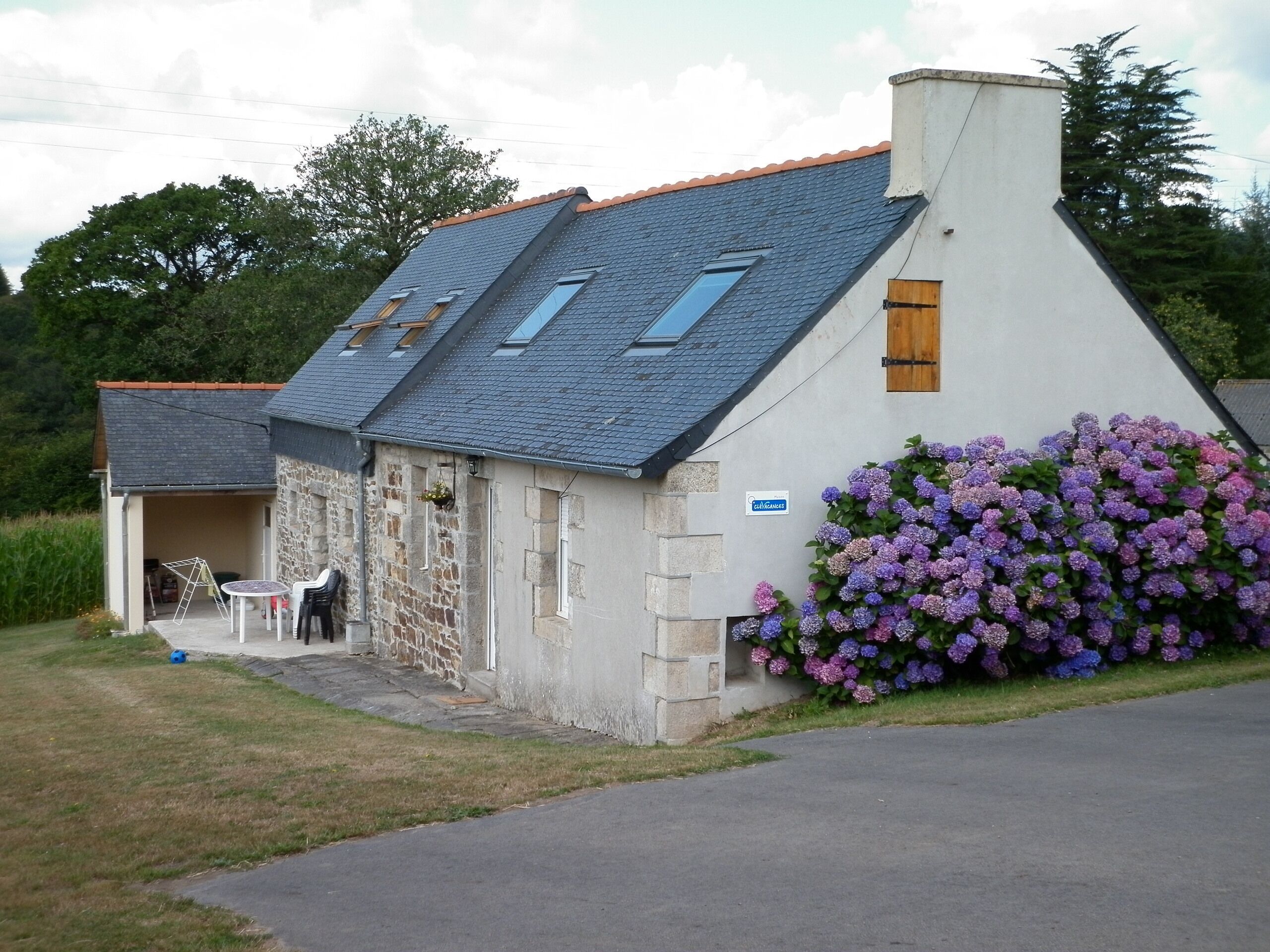 A stone building with a sloped roof, a small attached structure with a table and chairs, and a flower bed with purple flowers.