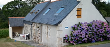 A stone building with a sloped roof, a small attached structure with a table and chairs, and a flower bed with purple flowers.