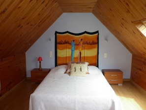 A bedroom with a sloped ceiling, wooden walls, a bed with a white bedspread, two bedside tables with lamps, and a colorful tapestry on the wall.