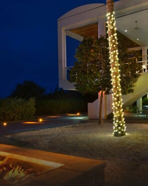 Un edificio moderno con fachada de vidrio, un árbol iluminado con luces y un pozo de fuego al frente.