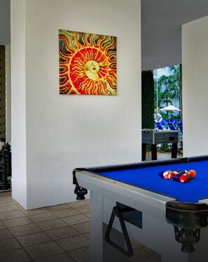 Sala de billar con mesa de pool, un cuadro llamativo de un sol y vistas a una piscina con sillones de descanso.