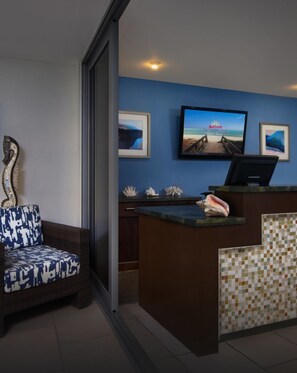 Un vestíbulo con mostrador de recepción, pared azul, televisor mostrando una escena de playa y una escultura decorativa de cisne.