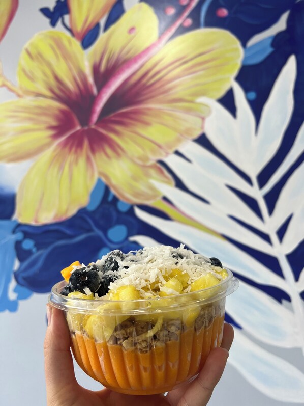Una mano sostiene un vaso de plástico transparente con un postre en capas, que incluye mango, coco y posiblemente otras frutas, sobre un fondo con una flor grande y hojas.