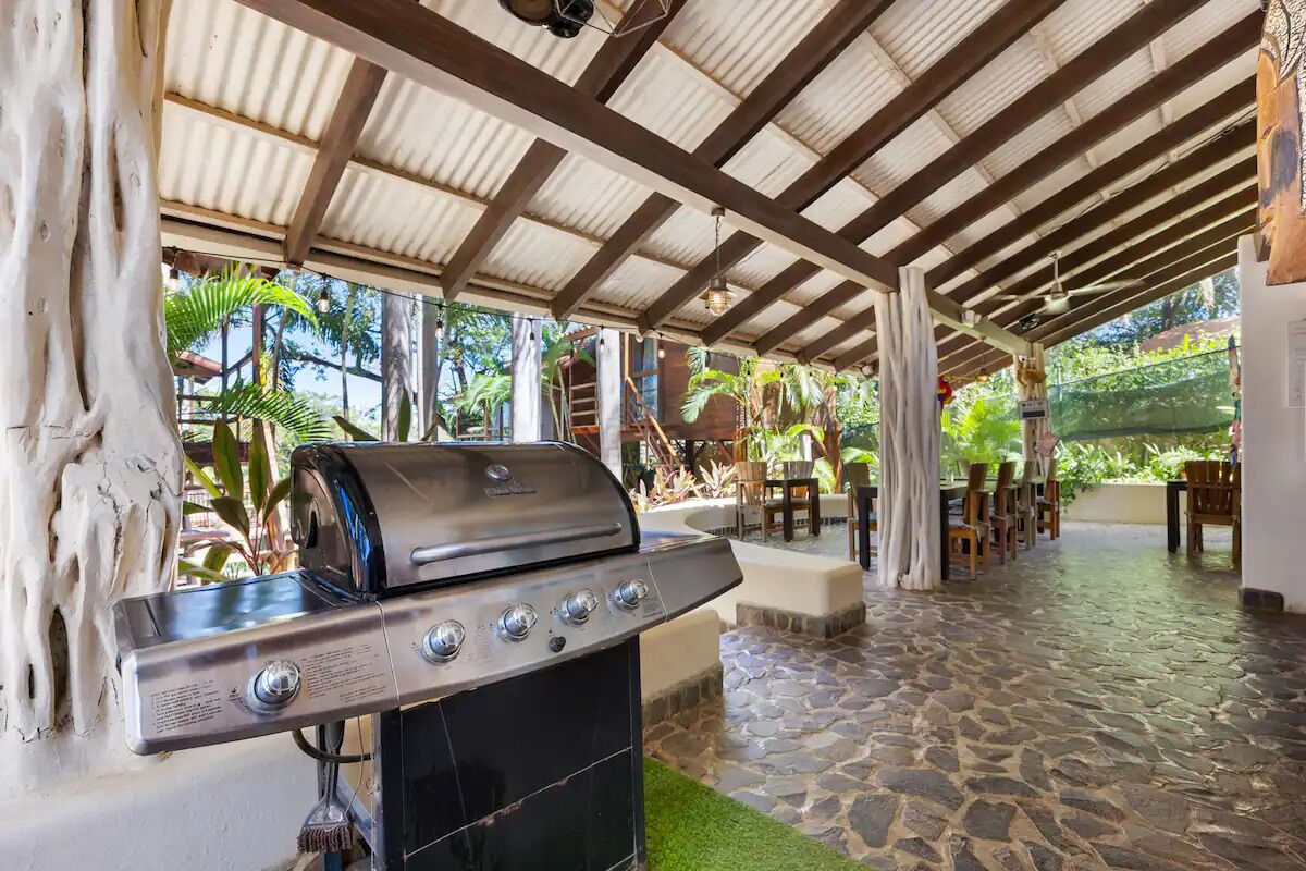 An outdoor covered patio area with a barbecue grill, stone flooring, and tropical plants.