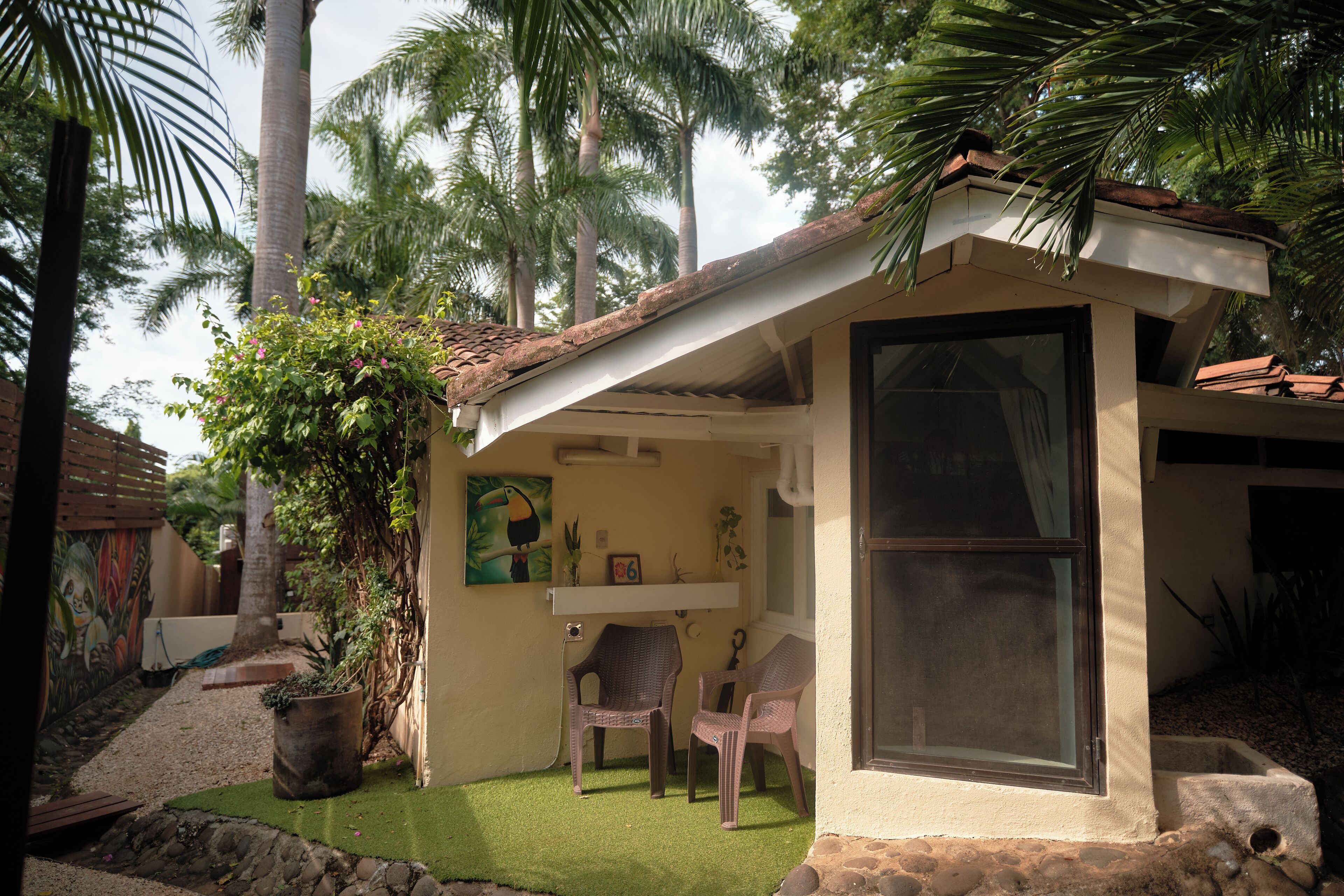 A small, single-story building with a sloped roof, a large window, and a door. There are two wicker chairs outside and a potted plant nearby.