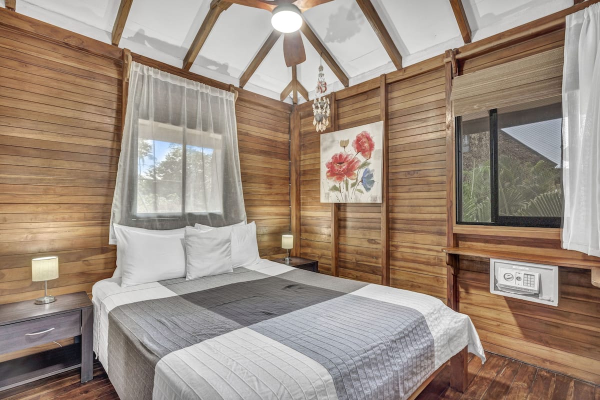 A wooden-paneled room with a bed, two bedside tables, a ceiling fan, a window with curtains, and a painting.
