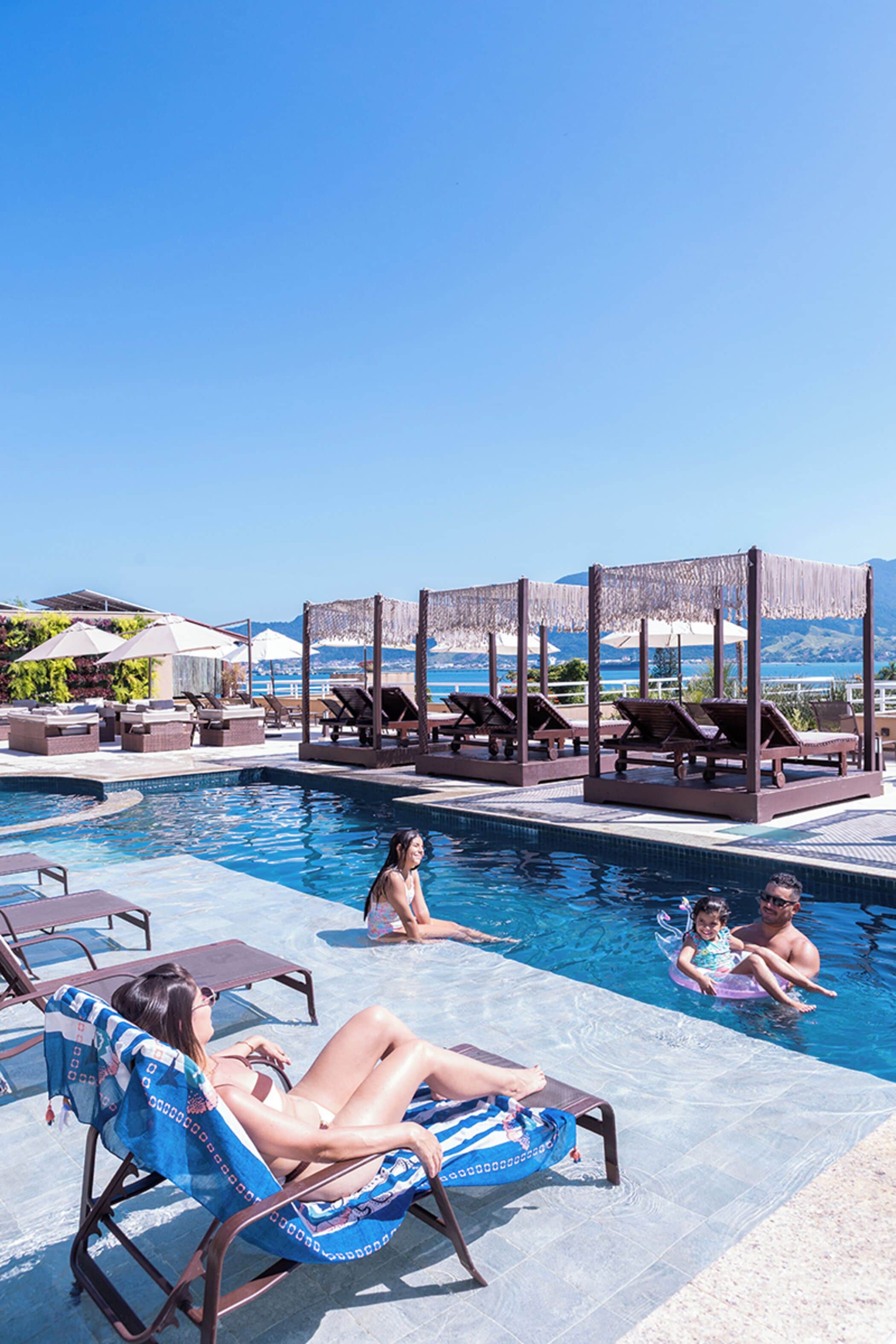 Zona de piscina con sillones de descanso y sombrillas, cielo despejado y vista al mar.