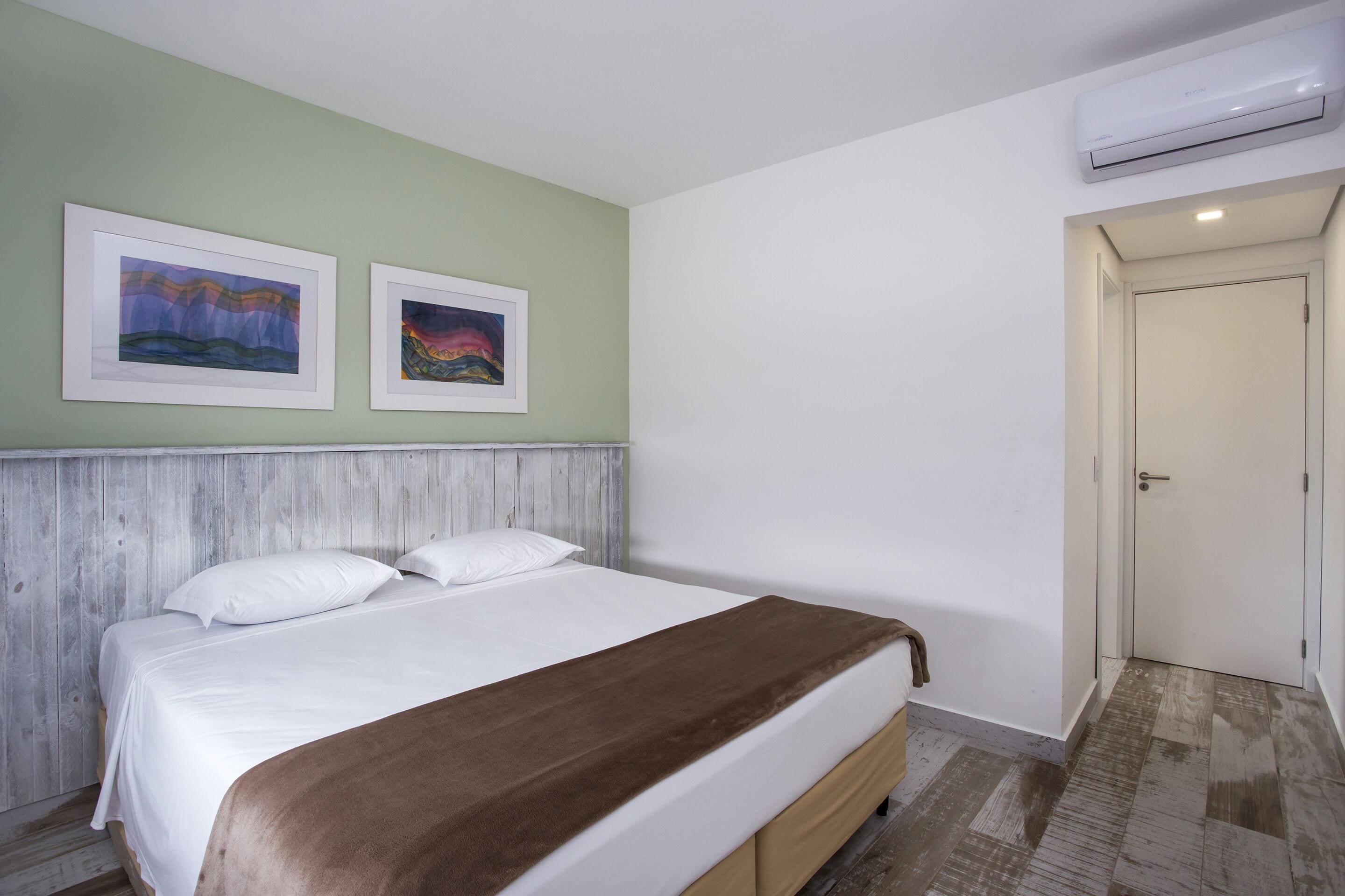 Une chambre à coucher avec un lit, du linge de lit blanc, un plaid marron, deux œuvres d’art encadrées accrochées au mur et un appareil de climatisation au-dessus de la porte.