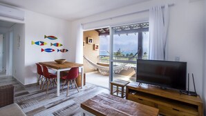 A modern living room with a wooden dining table, red chairs, a large flat-screen TV, and a hammock by a large window with a view. - Ilhabela Beach Residences (Ilhabela)