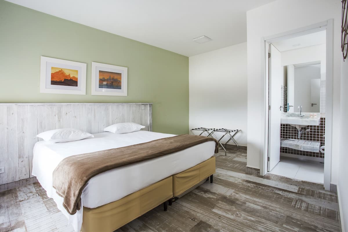 A neatly arranged hotel room with a bed, a wooden headboard, a green wall, and a bathroom visible through an open door.