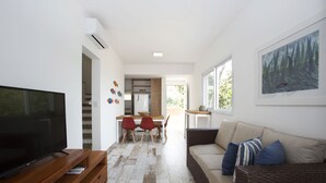 A modern living room with a flat-screen TV, a sofa with cushions, a dining area with chairs, and a kitchen in the background. - Ilhabela Beach Residences (Ilhabela)
