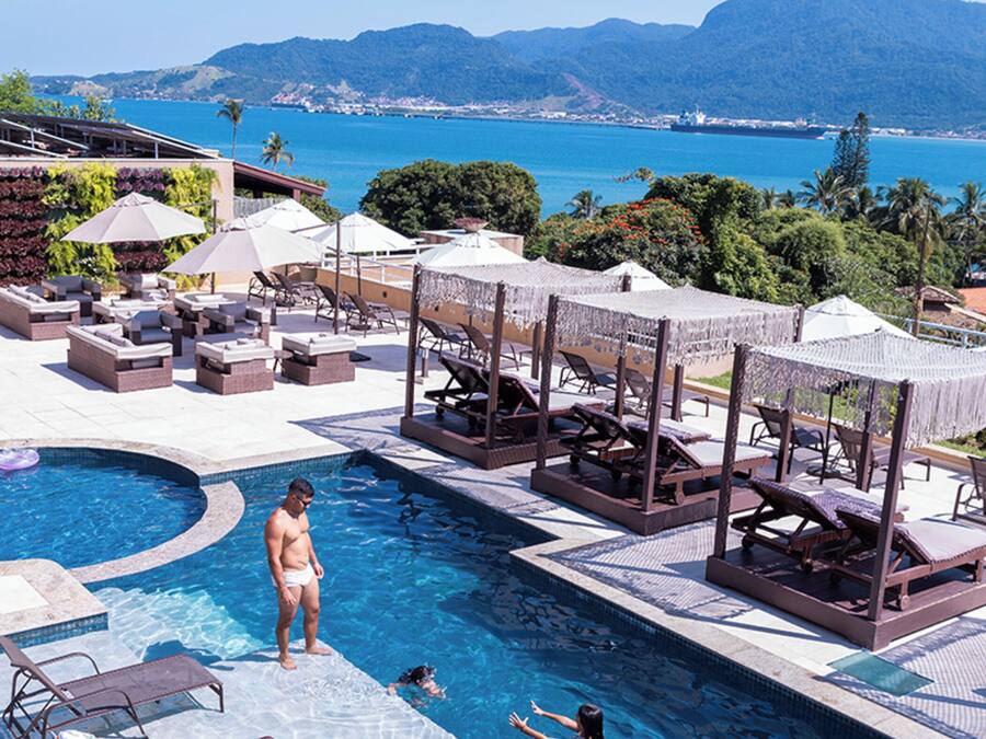 A pool area with lounge chairs and umbrellas, a swimming pool, and a view of the ocean and mountains.