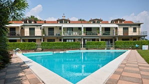 Ein rechteckiger Pool mit Fliesen umgeben von einer gepflegten Hecke und einem mehrstöckigen Gebäude mit Balkonen im Hintergrund.