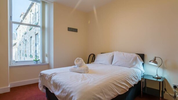 A neatly made bed with white linens and a rolled towel, a bedside lamp, and a window with a view of buildings. - Heart of Finnieston (Glasgow)