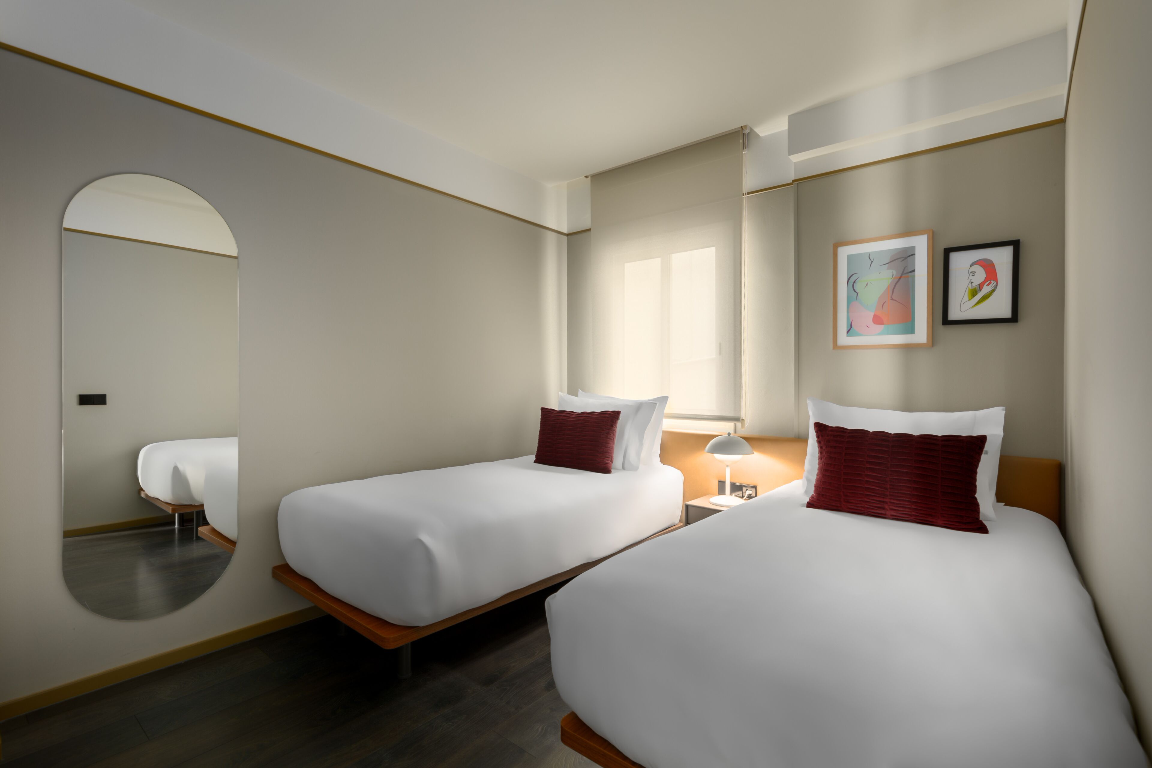 Une chambre d’hôtel avec deux lits simples, un grand miroir, une table de chevet avec une lampe et des œuvres d’art accrochées au mur.