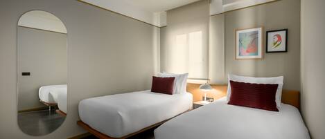 Une chambre d’hôtel avec deux lits simples, un grand miroir, une table de chevet avec une lampe et des œuvres d’art accrochées au mur.