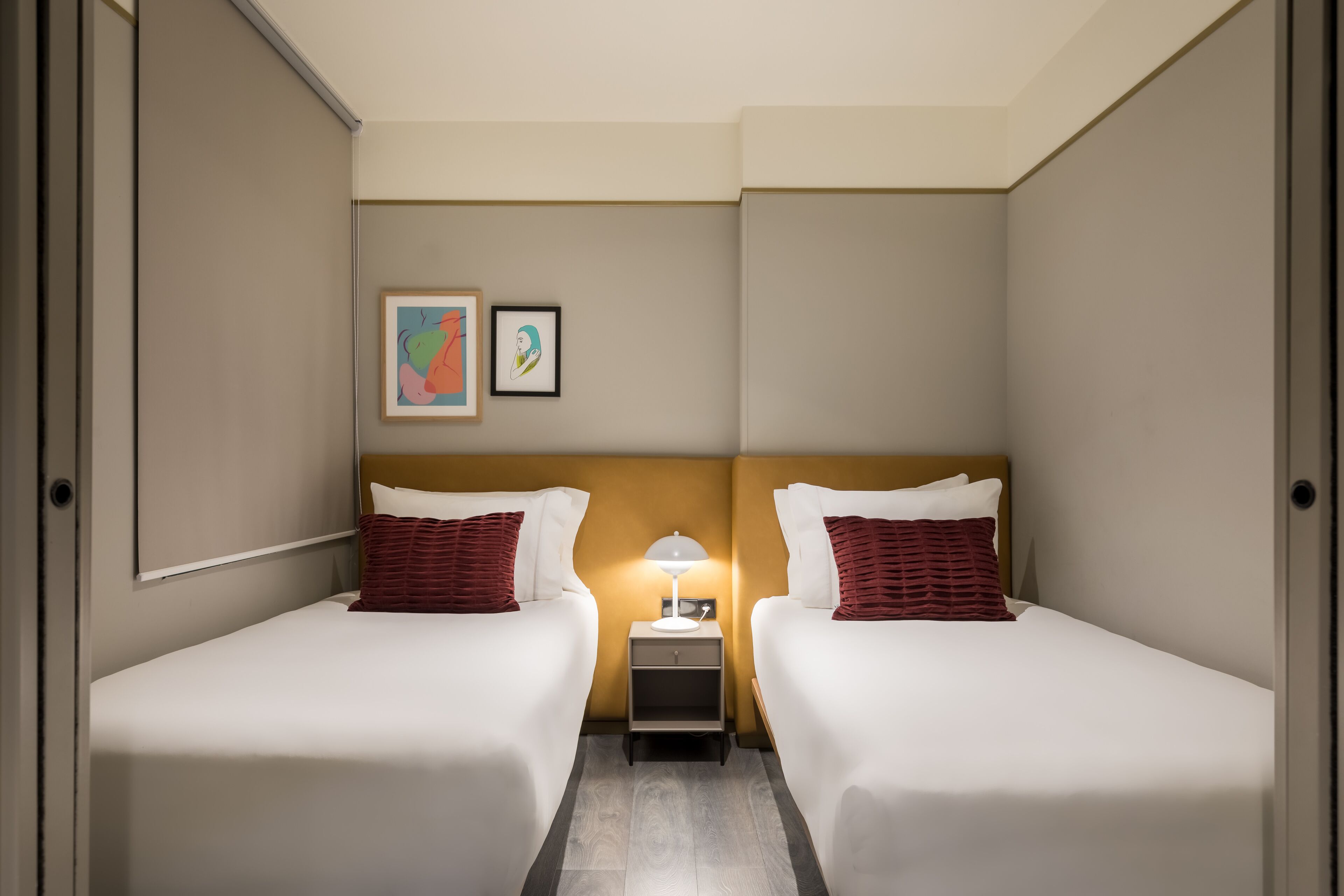 Habitación de hotel con dos camas, una mesita de noche, una lámpara y dos cuadros en la pared.