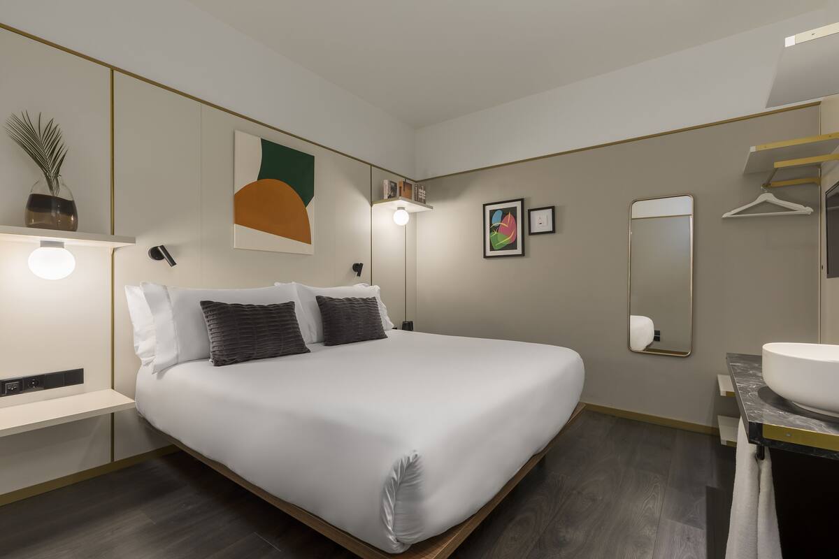 Ein modernes Hotelzimmer mit einem großen Bett, Nachttischen, einer Nachttischlampe, einem wandmontierten Spiegel und einem Badezimmer mit Waschbecken und Handtuchhalter.