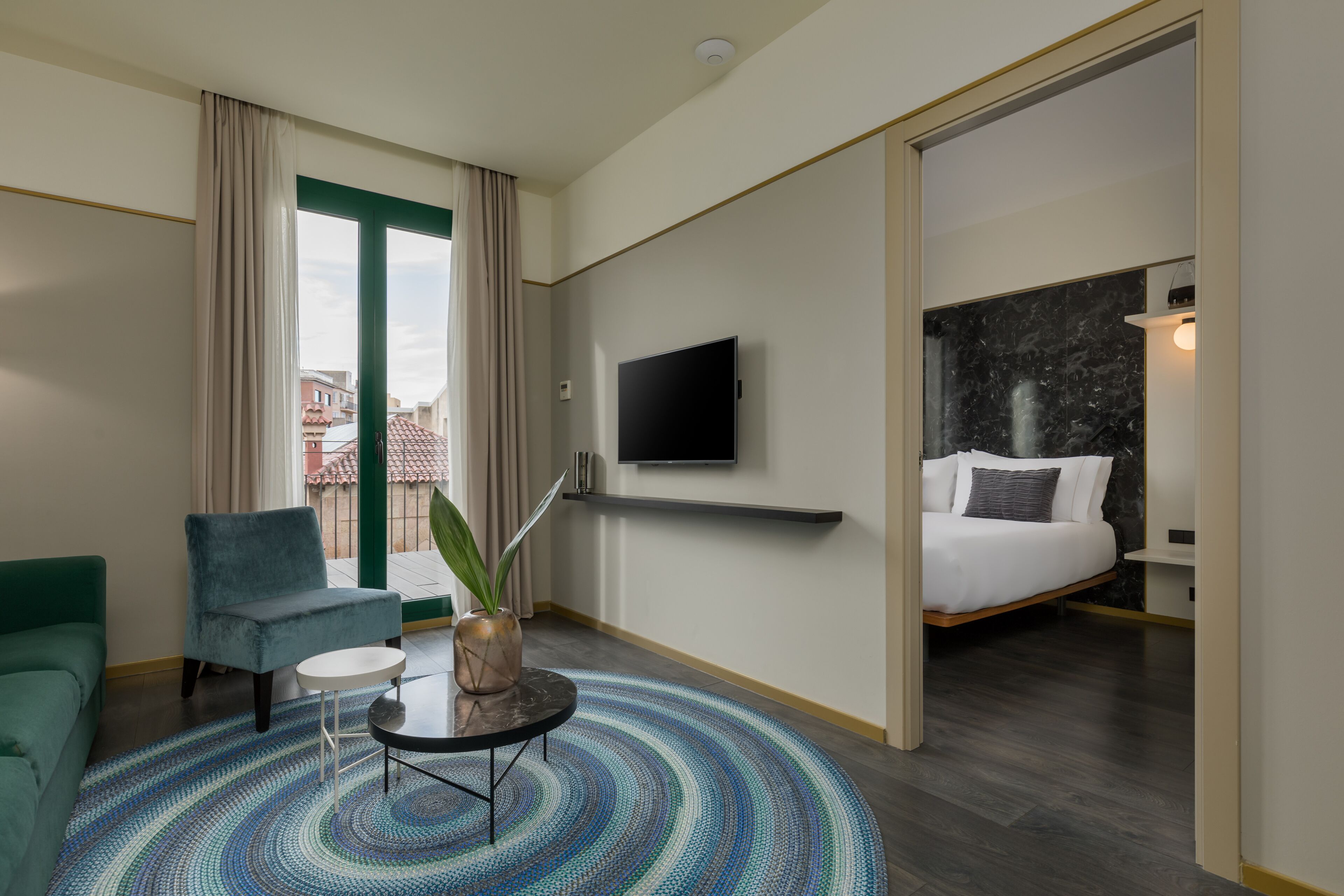 Una habitación de hotel con cama, televisor, un sillón verde, una pequeña mesa con una planta y vistas a la ciudad a través de un ventanal grande.