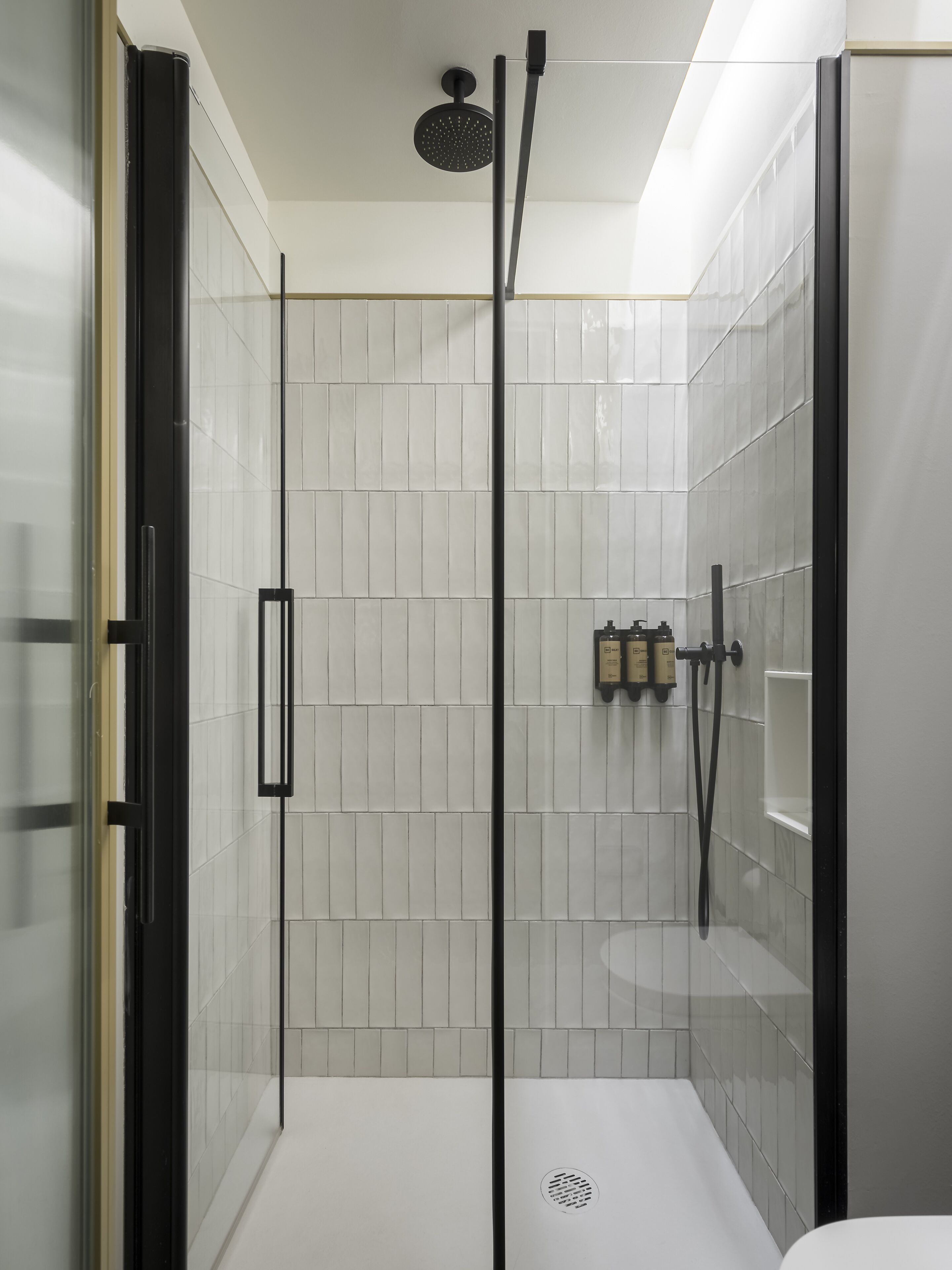 A modern shower area with a glass door, white tiled walls, and a ceiling-mounted showerhead.