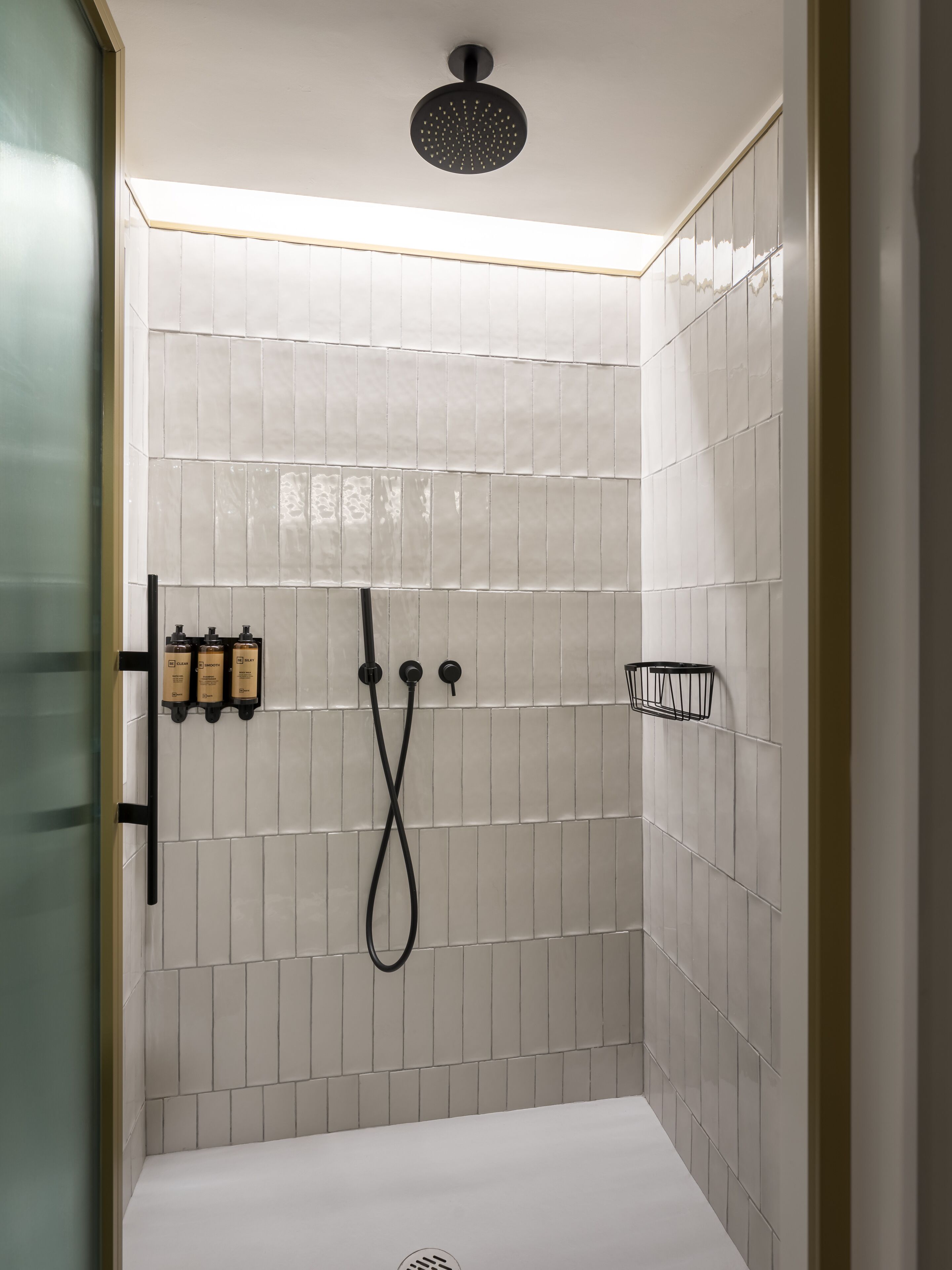 Ducha moderna con cabezal de ducha lluvia negro, un cabezal de ducha manual y otro cabezal de ducha manual con manguera negra. Hay tres botellas de champú y acondicionador en la pared.