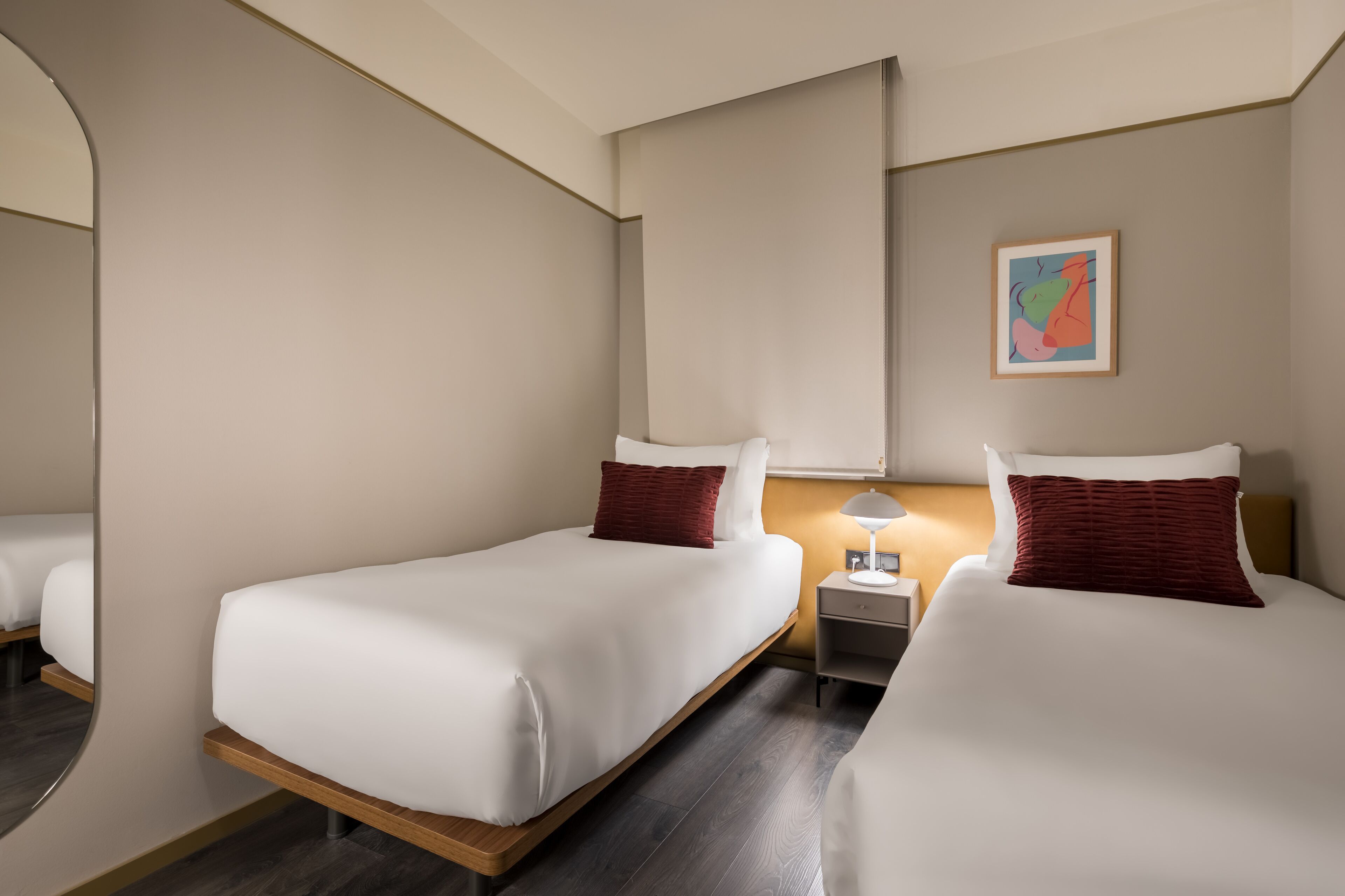 A hotel room with two single beds, a nightstand with a lamp, and a framed artwork on the wall.