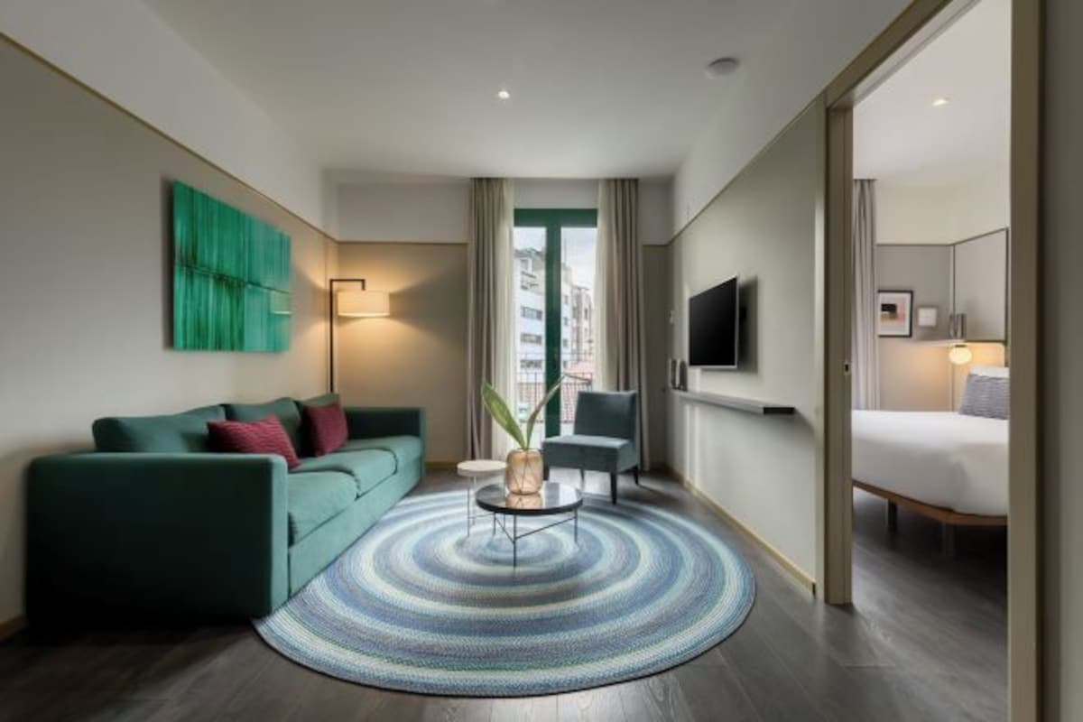 A modern hotel room with a green sofa, a round coffee table, a TV, and a bed.