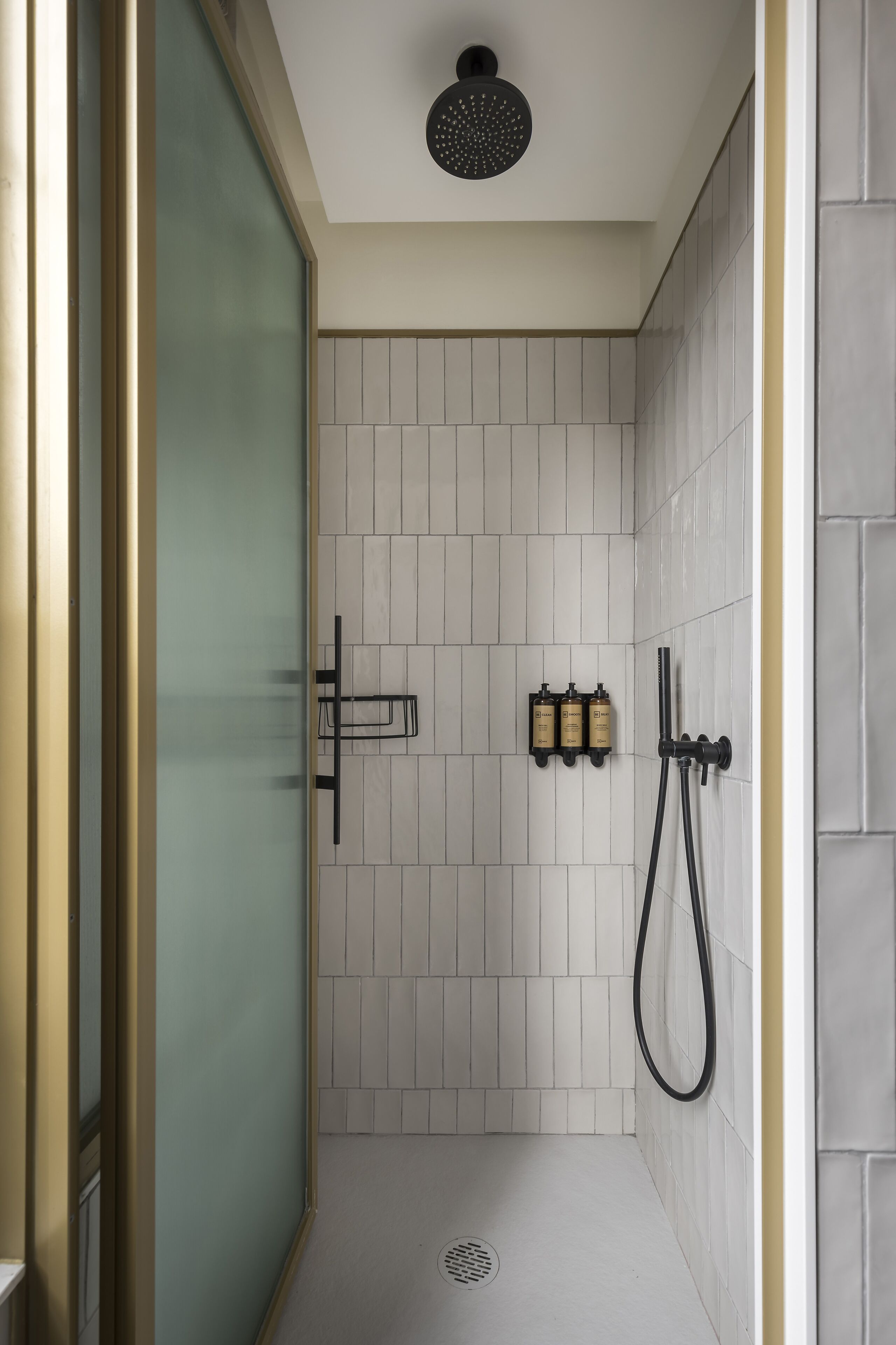 A modern shower with a glass door, white tiled walls, and a black showerhead.