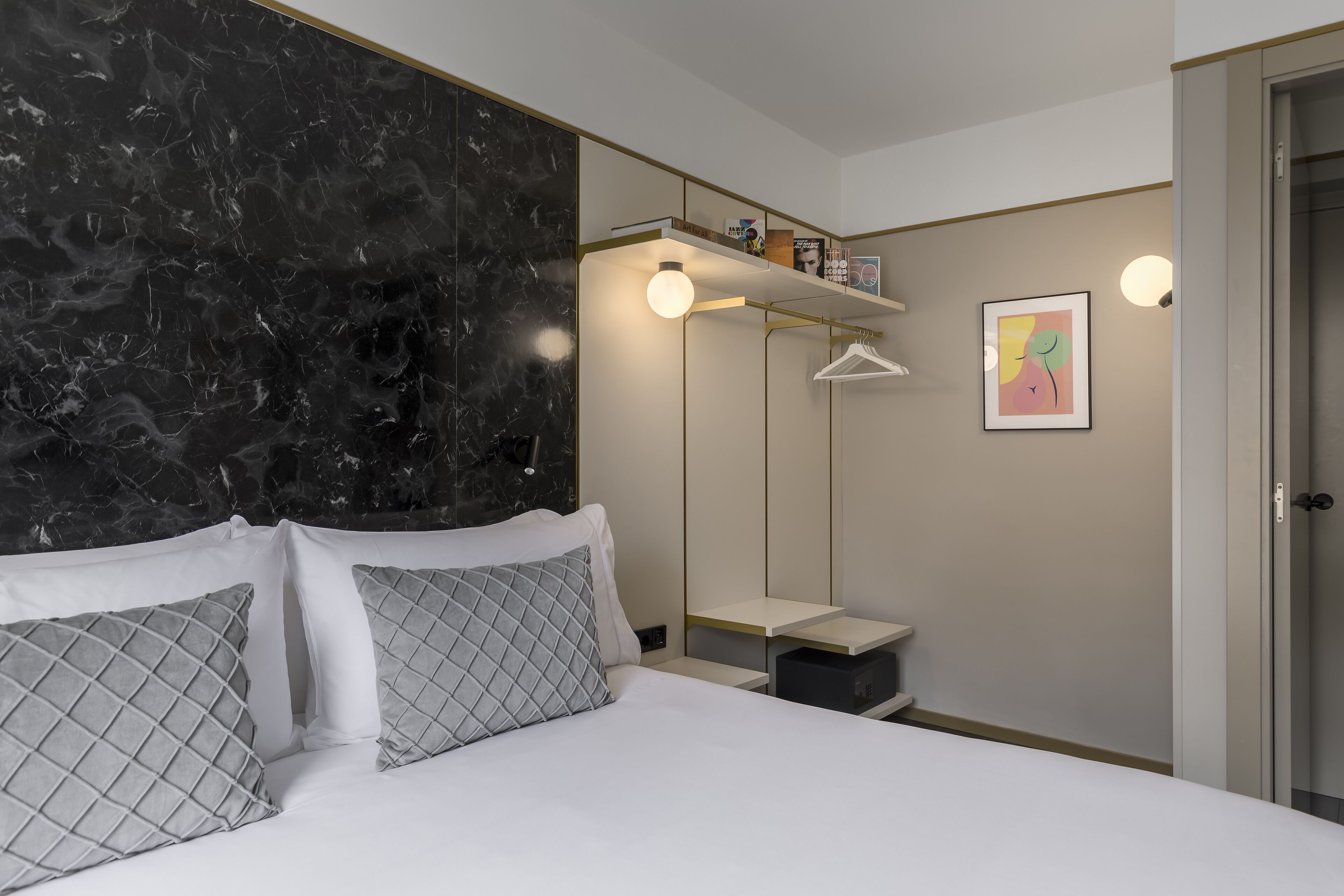 Habitación de hotel moderna con cabecera de mármol, cama con sábanas blancas y almohadones estampados, mesita de noche con lámpara, armario y una pequeña estantería con libros y objetos decorativos.