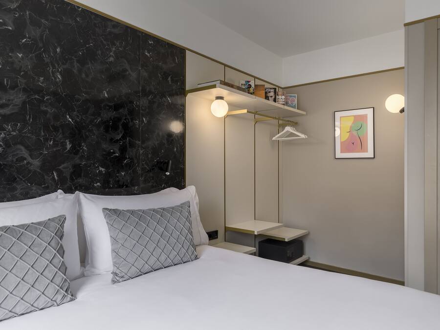 Habitación de hotel moderna con cabecera de mármol, cama con sábanas blancas y almohadones estampados, mesita de noche con lámpara, armario y una pequeña estantería con libros y objetos decorativos.