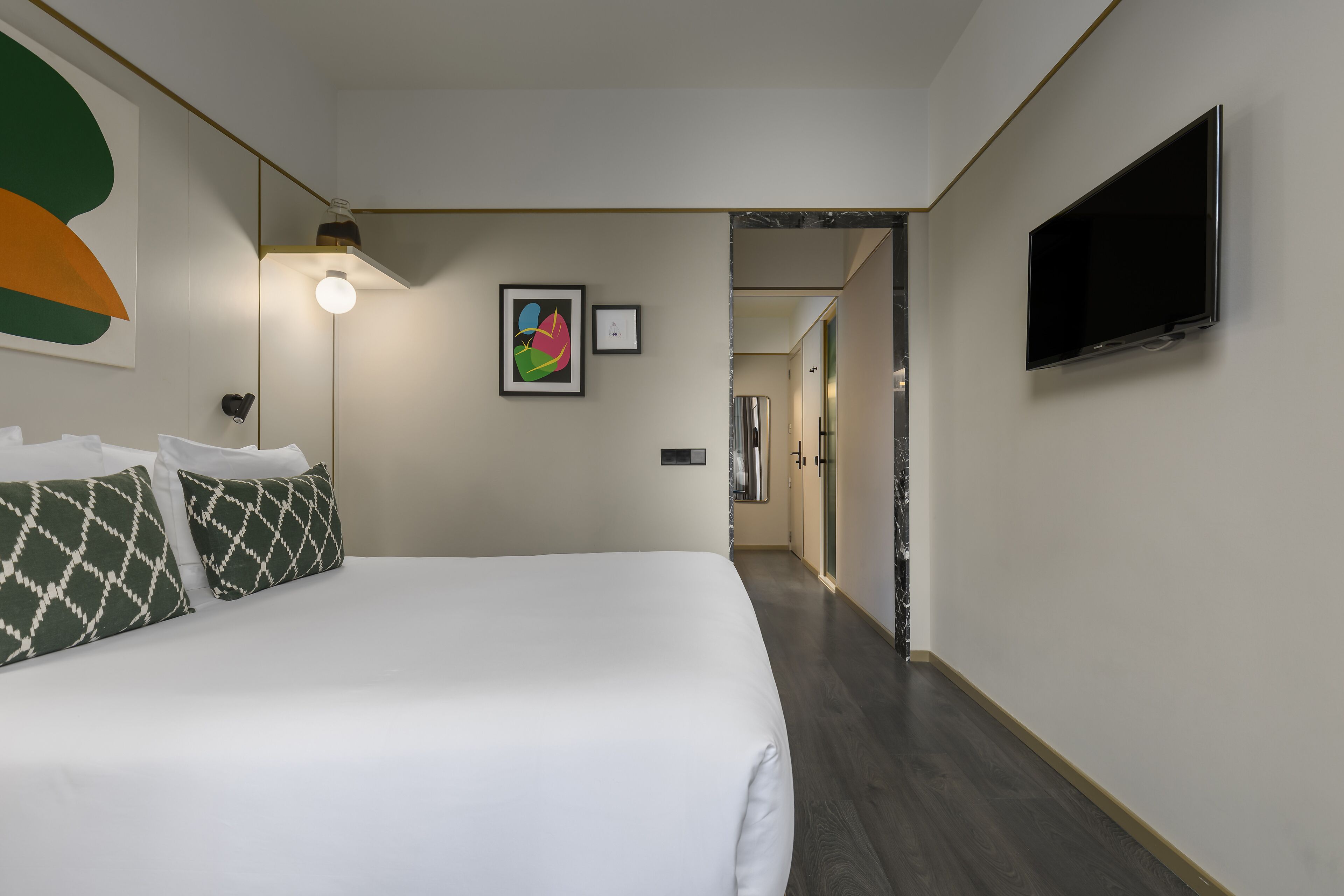 A hotel room with a large bed, a flat-screen TV mounted on the wall, and a hallway leading to other rooms.