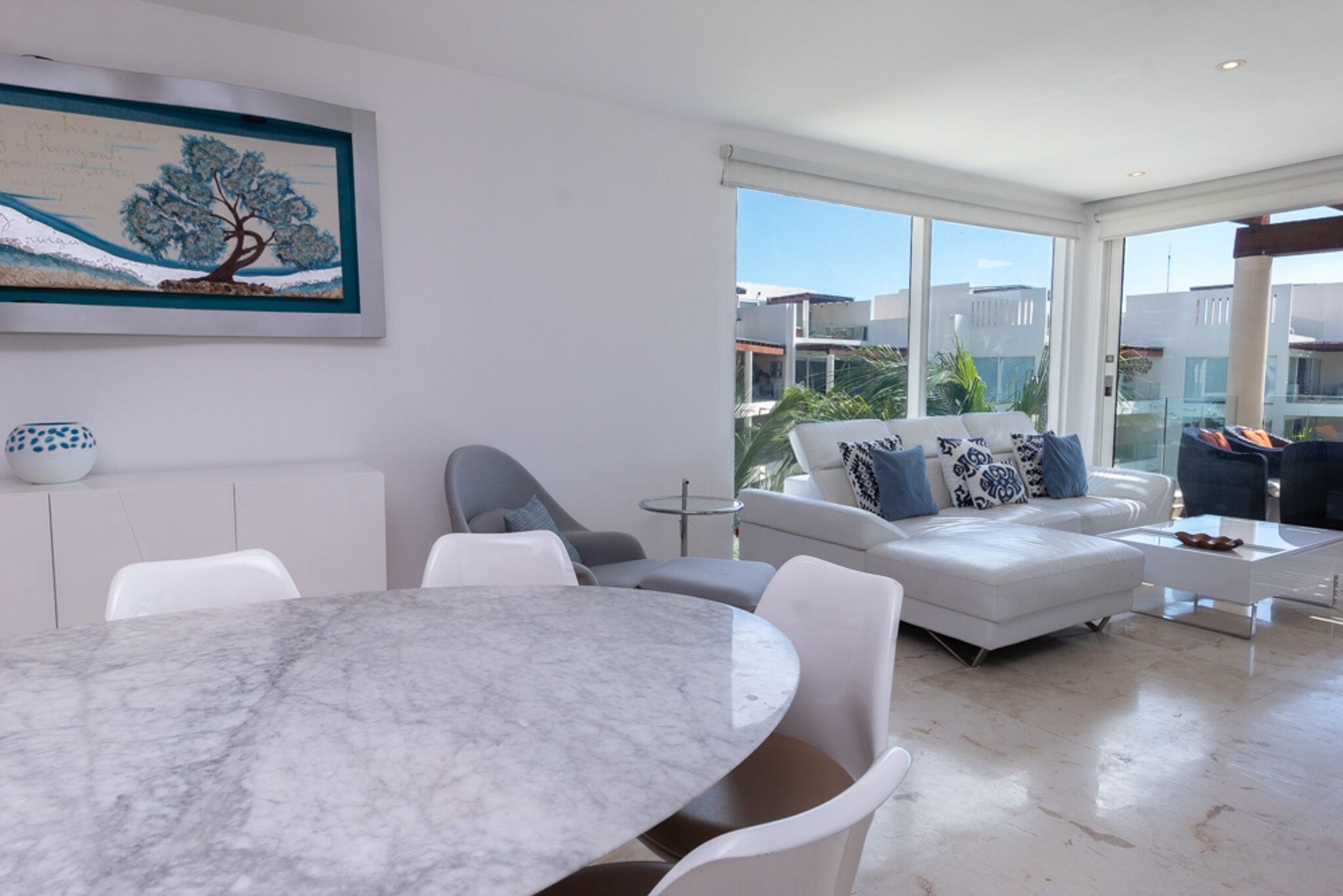 A modern living room with a marble table, white sofas, and a large window offering a view of buildings and greenery.