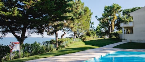 A swimming pool with a clear view of the sea, surrounded by lush greenery and a paved pathway.