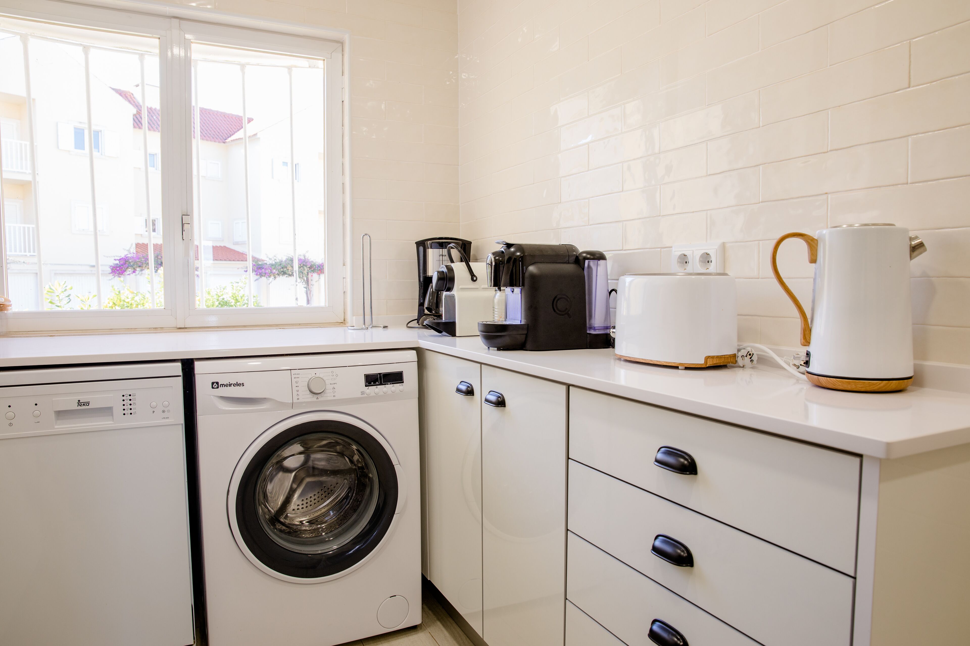 A compact kitchen with a washing machine, coffee maker, and kettle.