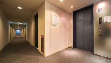 A hotel hallway with carpeted floors, a fire exit sign, an elevator with a digital display, and a door marked with the number 9.