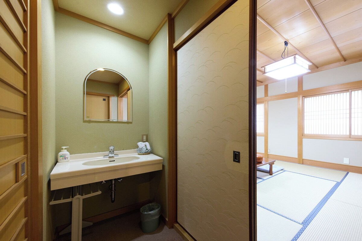 A traditional Japanese room with a tatami mat floor, a wooden ceiling, and a sliding door leading to a bathroom with a sink and mirror.