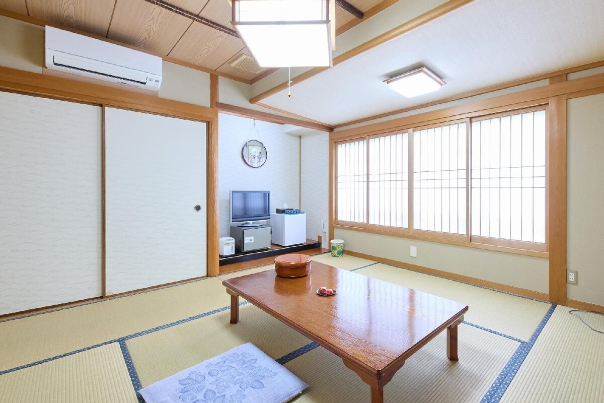 Una habitación japonesa tradicional con suelo de tatami, una mesa de madera, un televisor y una ventana con puertas shoji.