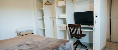 Ein Schlafzimmer mit einem Bett, einem Schreibtisch mit Stuhl, einem Fernseher und einem Kleiderschrank mit Regalen.