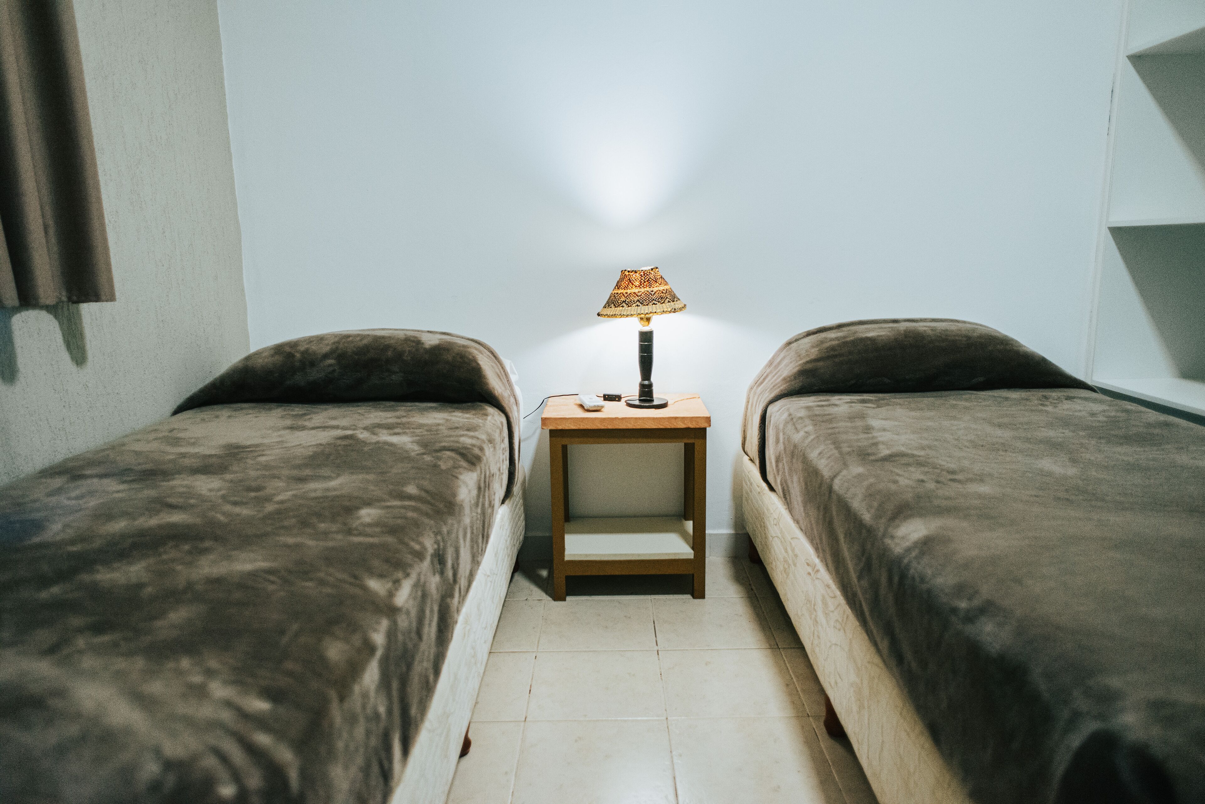 Alojamiento pequeño de una sola habitación con dos camas individuales, una mesita de noche con lámpara y una pared blanca lisa.
