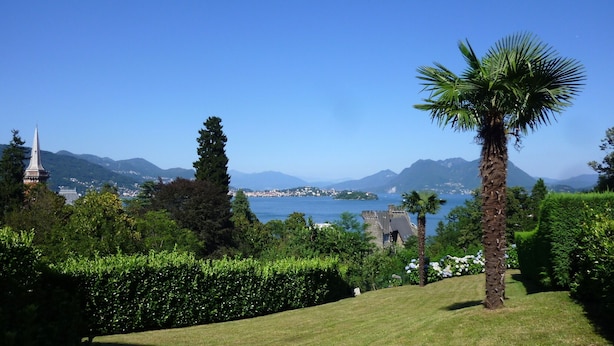 Een weelderige tuin met een palmboom, een kerktoren en een verre vergezichten over water en bergen.