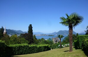 A lush garden with a palm tree, a church spire, and a distant view of a body of water and mountains. - VISTA CASTELLO - Apartment with lake and panoramic views + private garage! (Baveno)