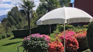 Een terras met een groene opklapstoel, een witte parasol en diverse kleurrijke bloemen en struiken.