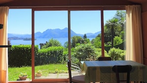 Een kamer met een groot raam, met uitzicht op een meer en bergen. Er staat een tafel met tafelkleed en stoelen.